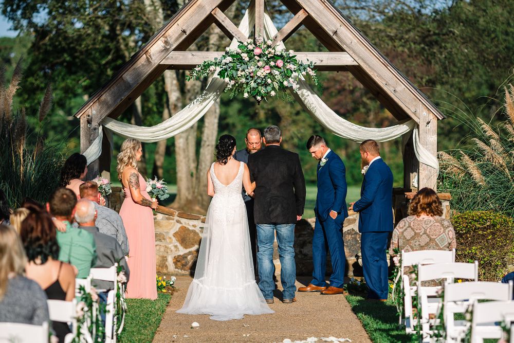 Outdoor wedding ceremony under decorated wooden arch with floral arrangements and white draping.
