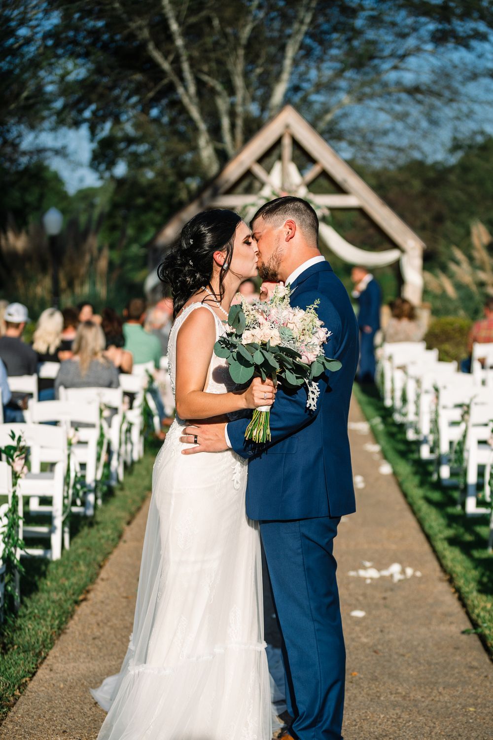 Romantic wedding kiss under rustic wooden arch with white floral arrangements.