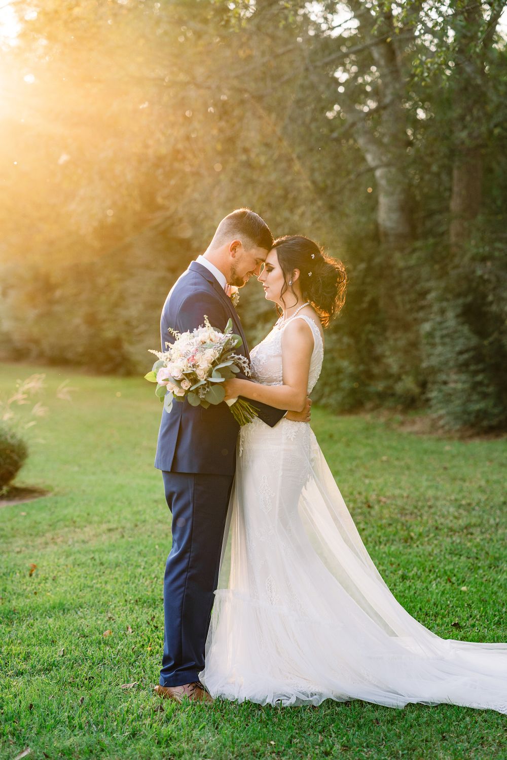 Romantic sunset wedding portrait with flowing white dress and pastel bouquet in garden setting.