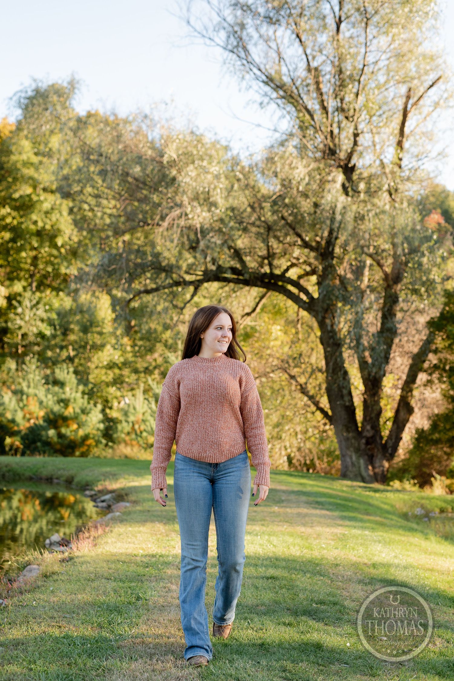 Person wearing pink sweater and jeans walks on grassy path with autumn trees in background.