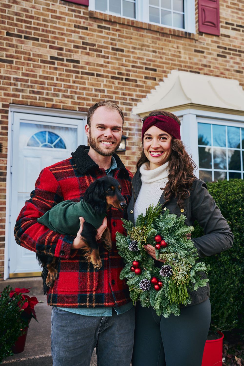 A couple poses outside their brick home with a holiday wreath and small black dog in festive winter attire.