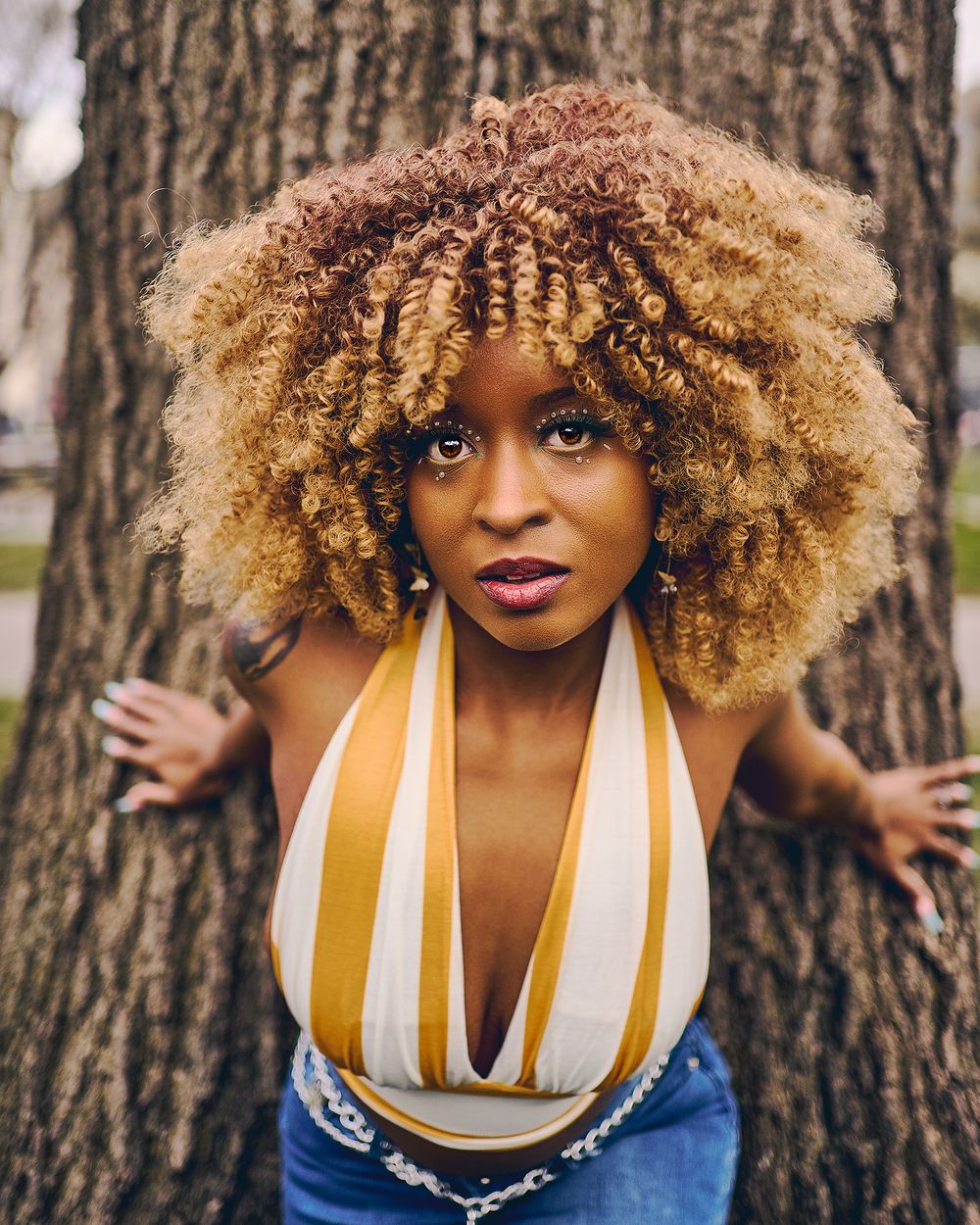 Natural portrait against tree trunk with curly highlighted hair and striped top.