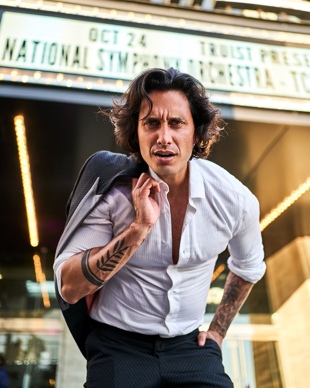 Person in white shirt with tattoos posing dramatically outside theater.