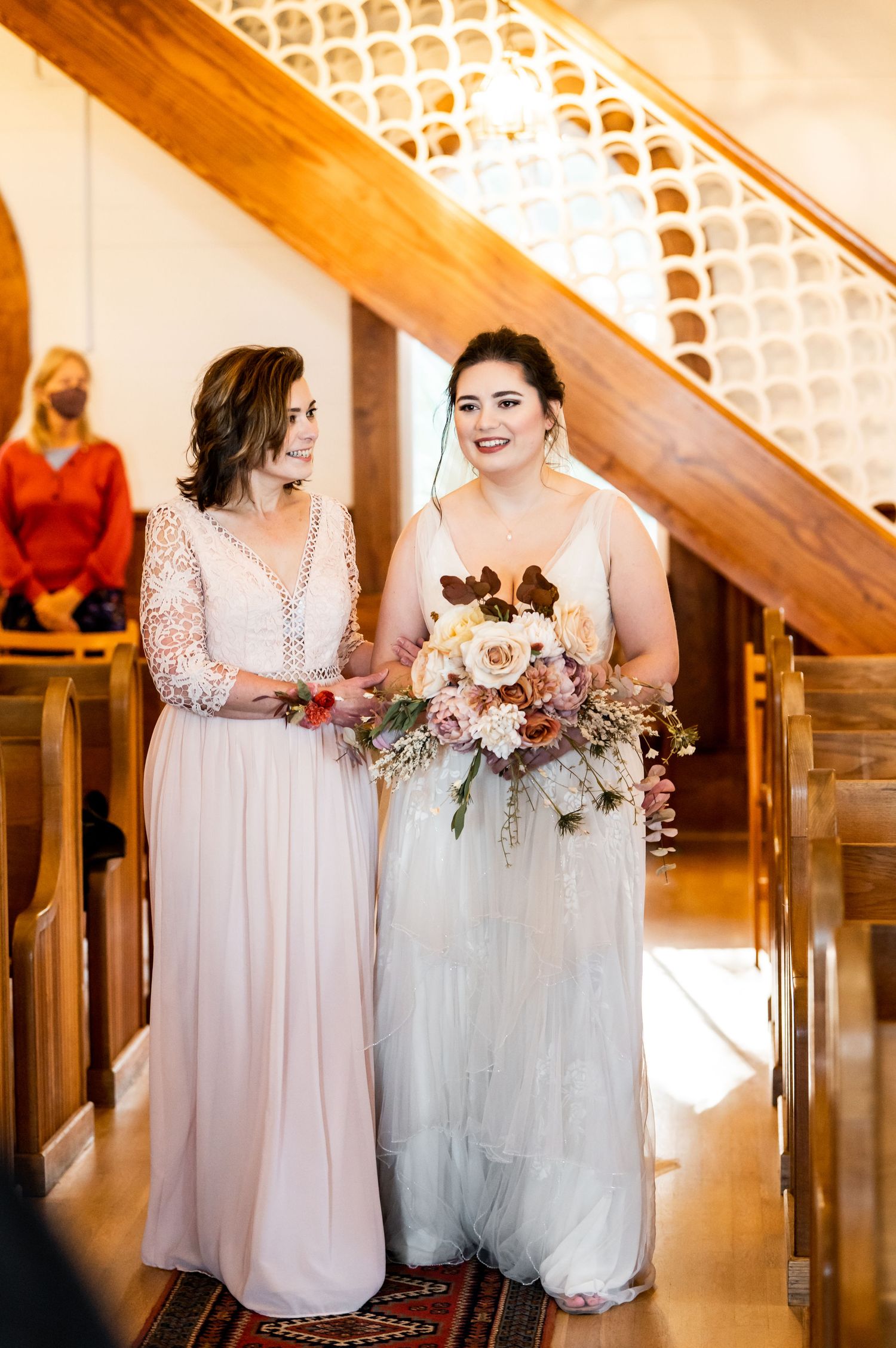 Two attendants in flowing pink dresses stand together holding bouquets during a wedding ceremony.