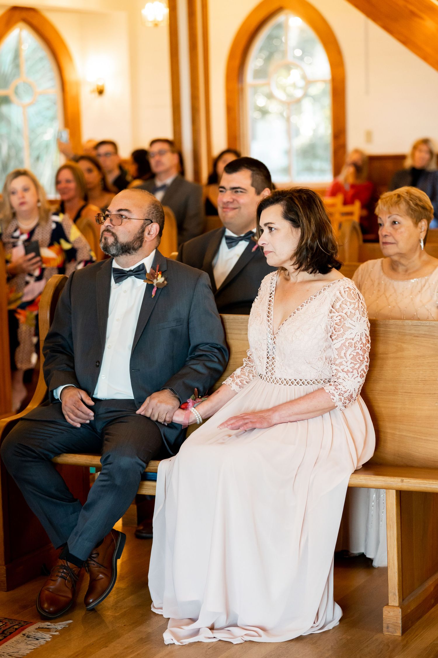 Wedding guests seated in wooden pews during ceremony in chapel with Gothic arched windows.
