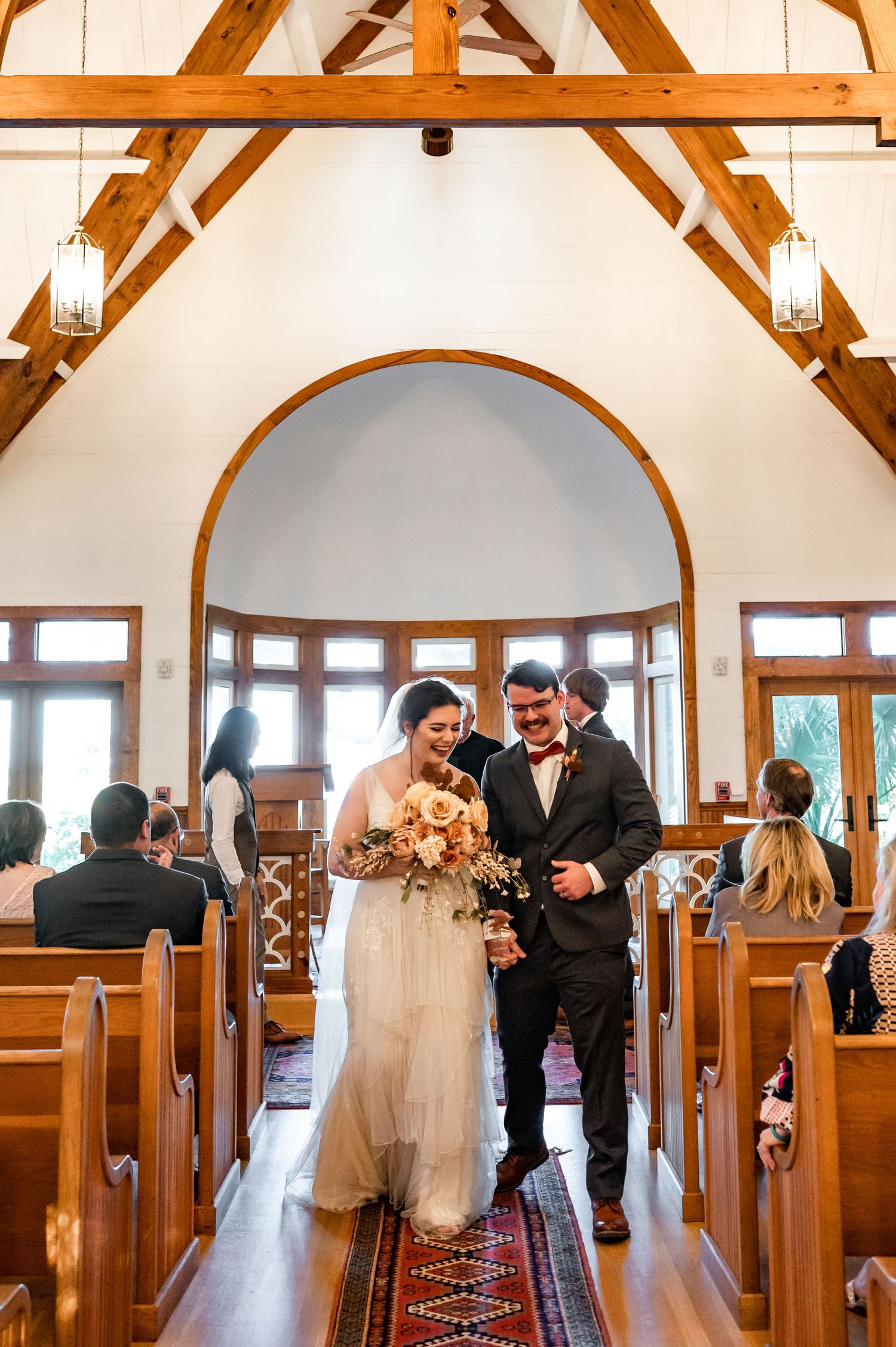 Wedding couple walks down aisle in chapel with vaulted wooden ceiling and stained glass window.