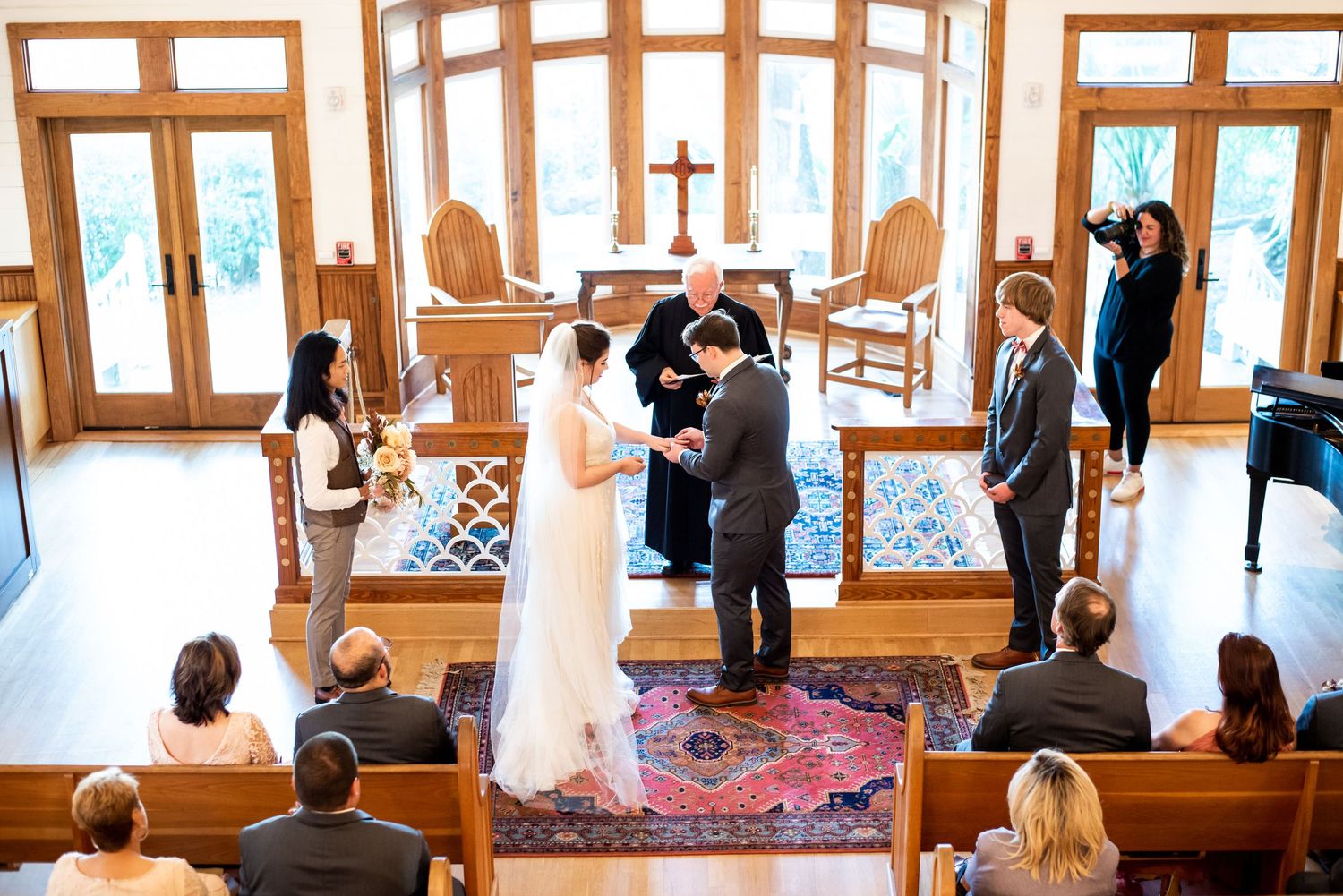 Wedding ceremony taking place in a bright chapel with wooden beams and large windows.
