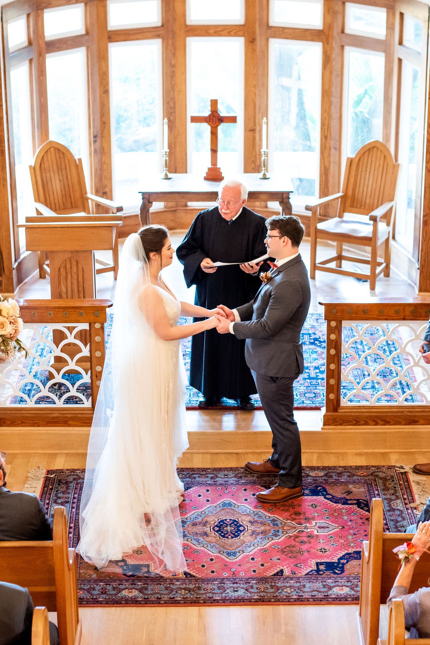 Wedding ceremony in progress inside a sunlit chapel with wooden details and oriental rug.