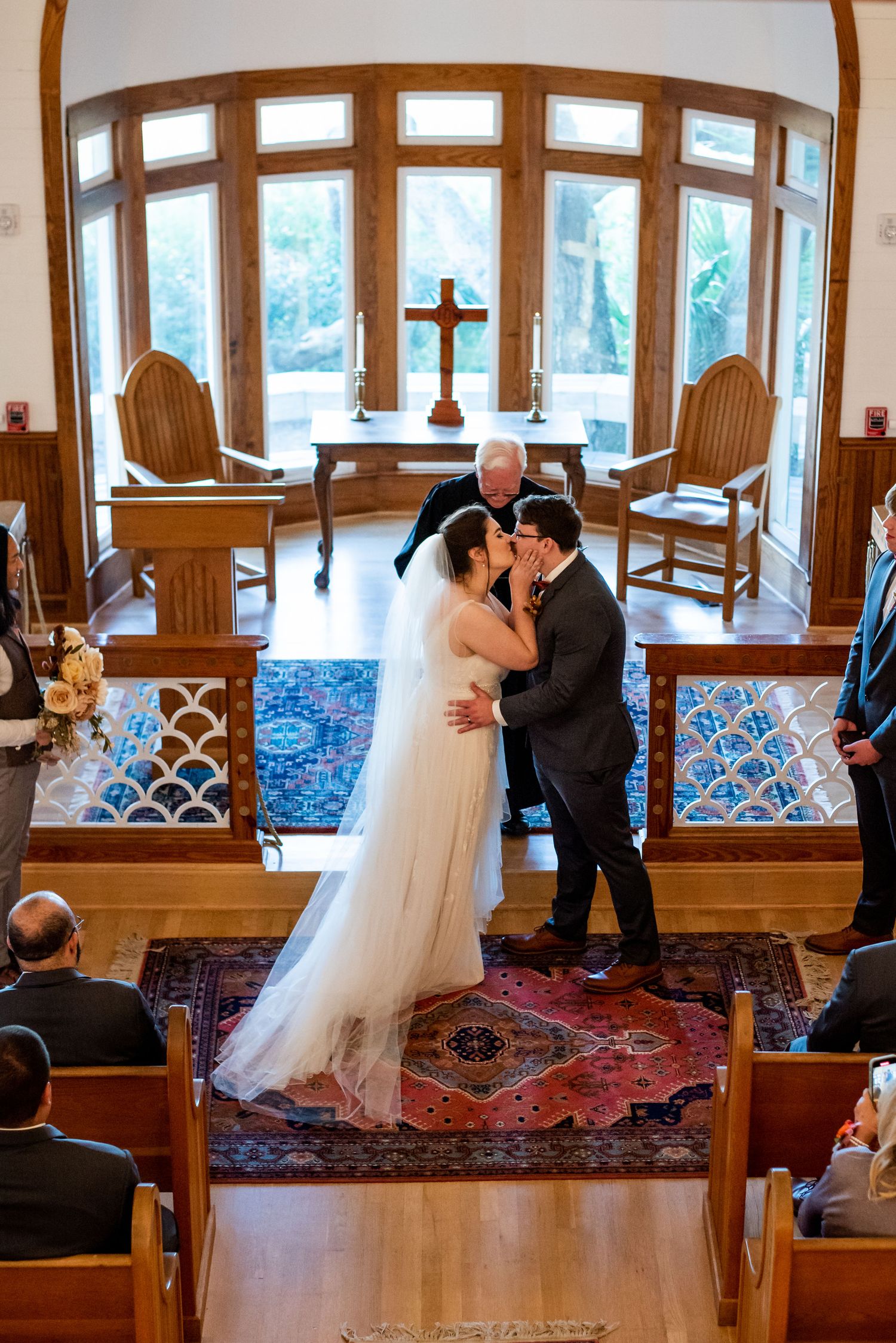 Romantic wedding ceremony moment in a chapel with large windows and wooden architectural details.