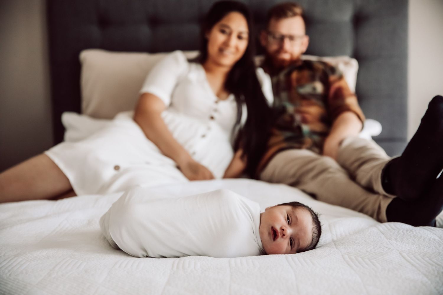 New parents lying in bed watching their swaddled newborn baby on white bedding.