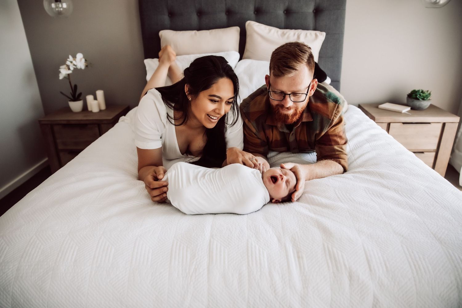 Couple lying on white bed admiring their swaddled newborn baby.