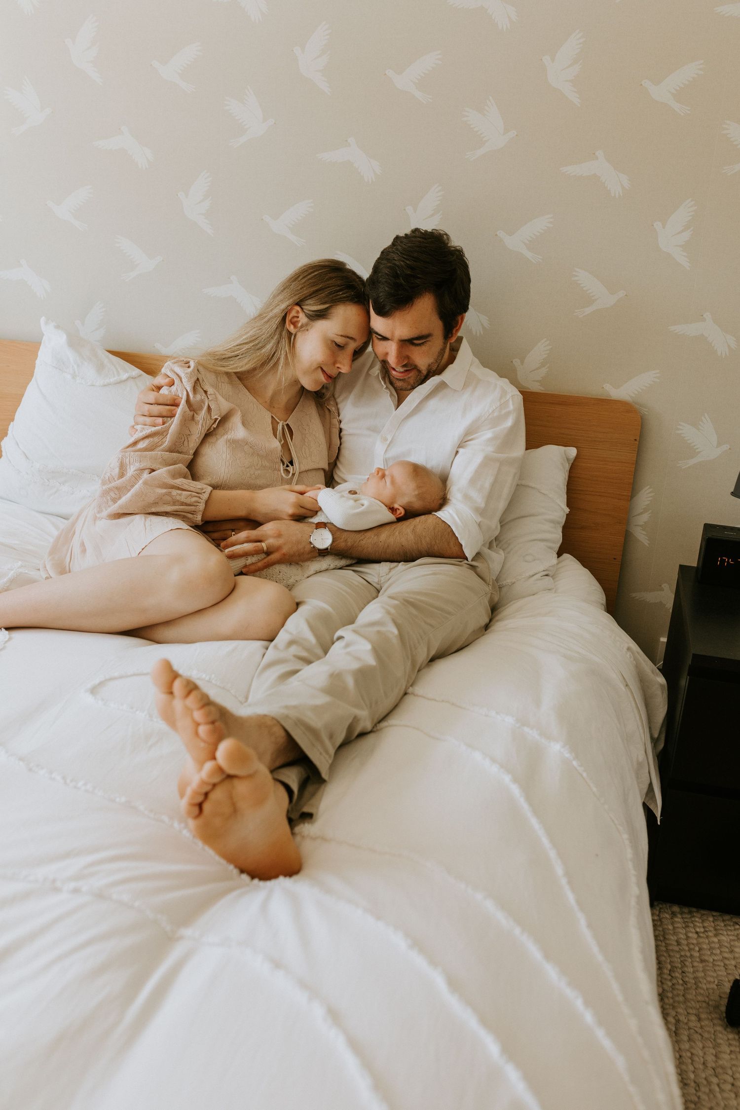 Tender family moments photographed on bed with bird wallpaper background in soft lighting.