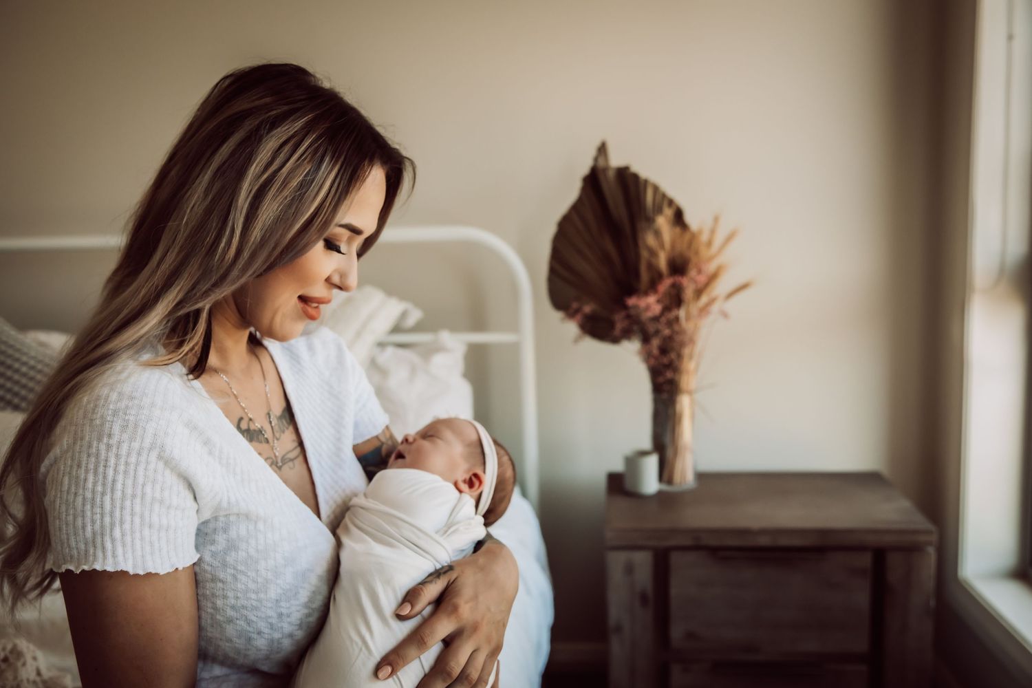 Mother in white sweater holding newborn baby near window with dried flowers.
