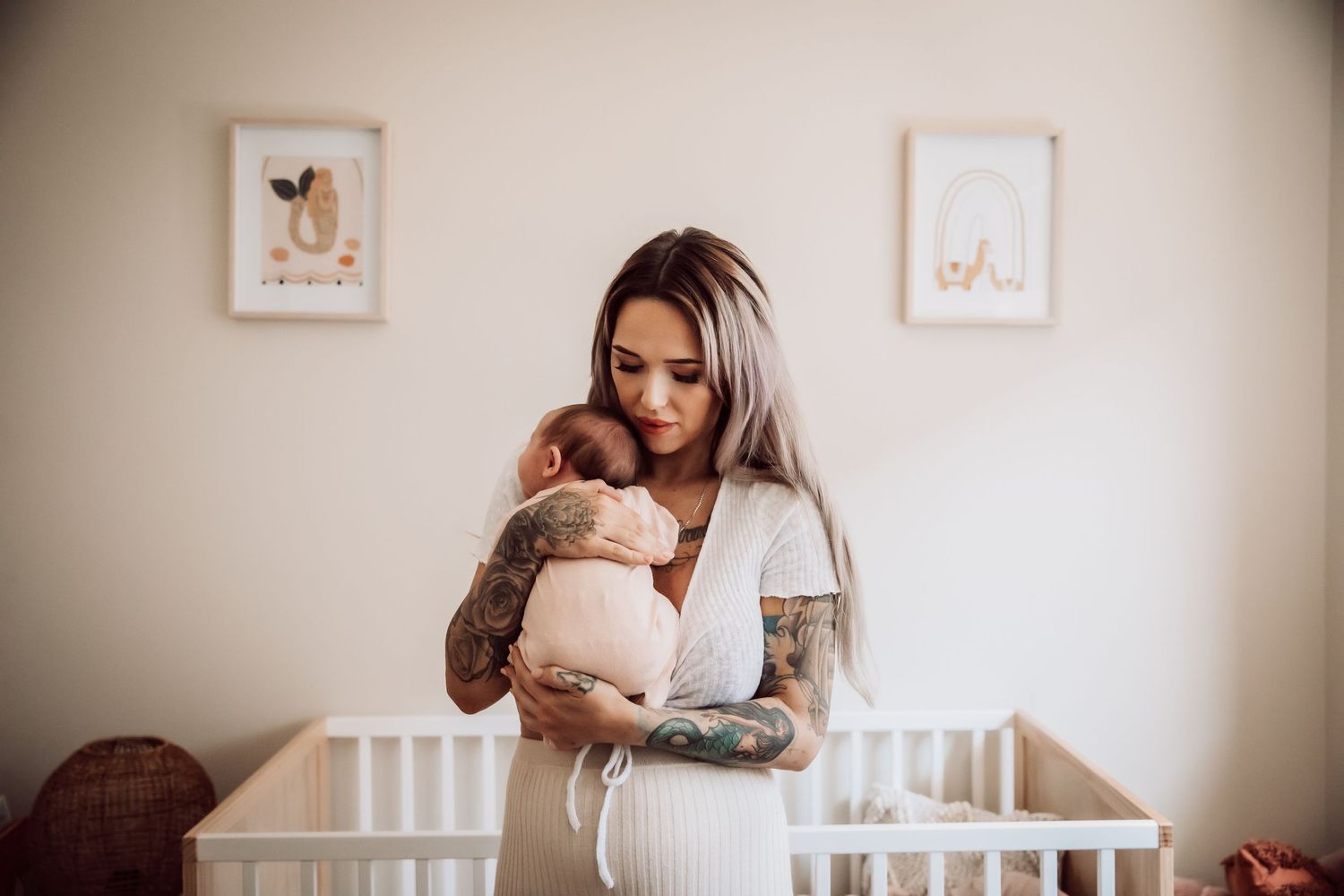 Mother with tattoos holding sleeping newborn in nursery with framed artwork.