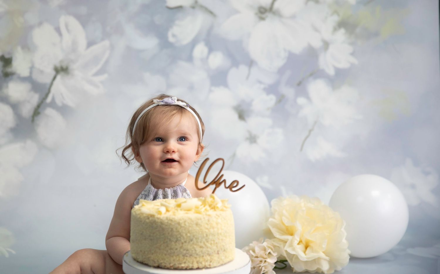 One year Cake Smash, baby girl wearing lilac lace bodysuit, which chocolate cake on stand, white balloons and cream pom