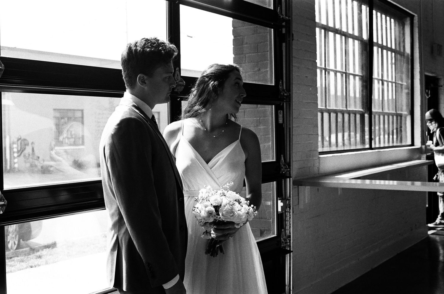 A couple shares an intimate moment in formal attire by large windows in black and white.