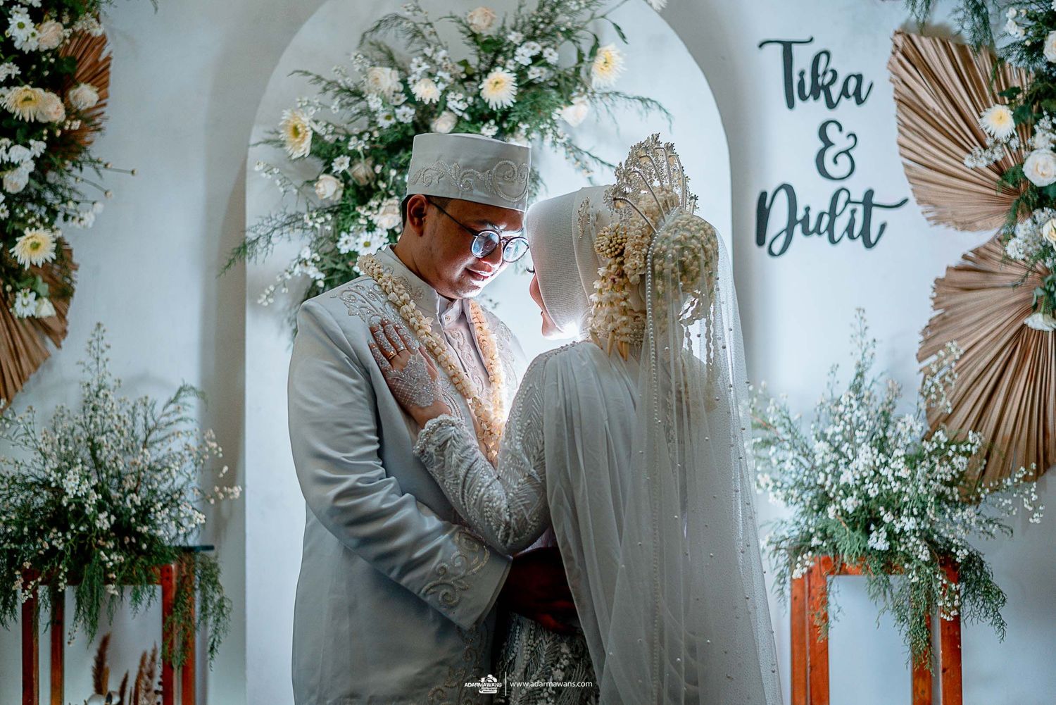 Romantic wedding moment under white archway decorated with flowers and palm leaves.