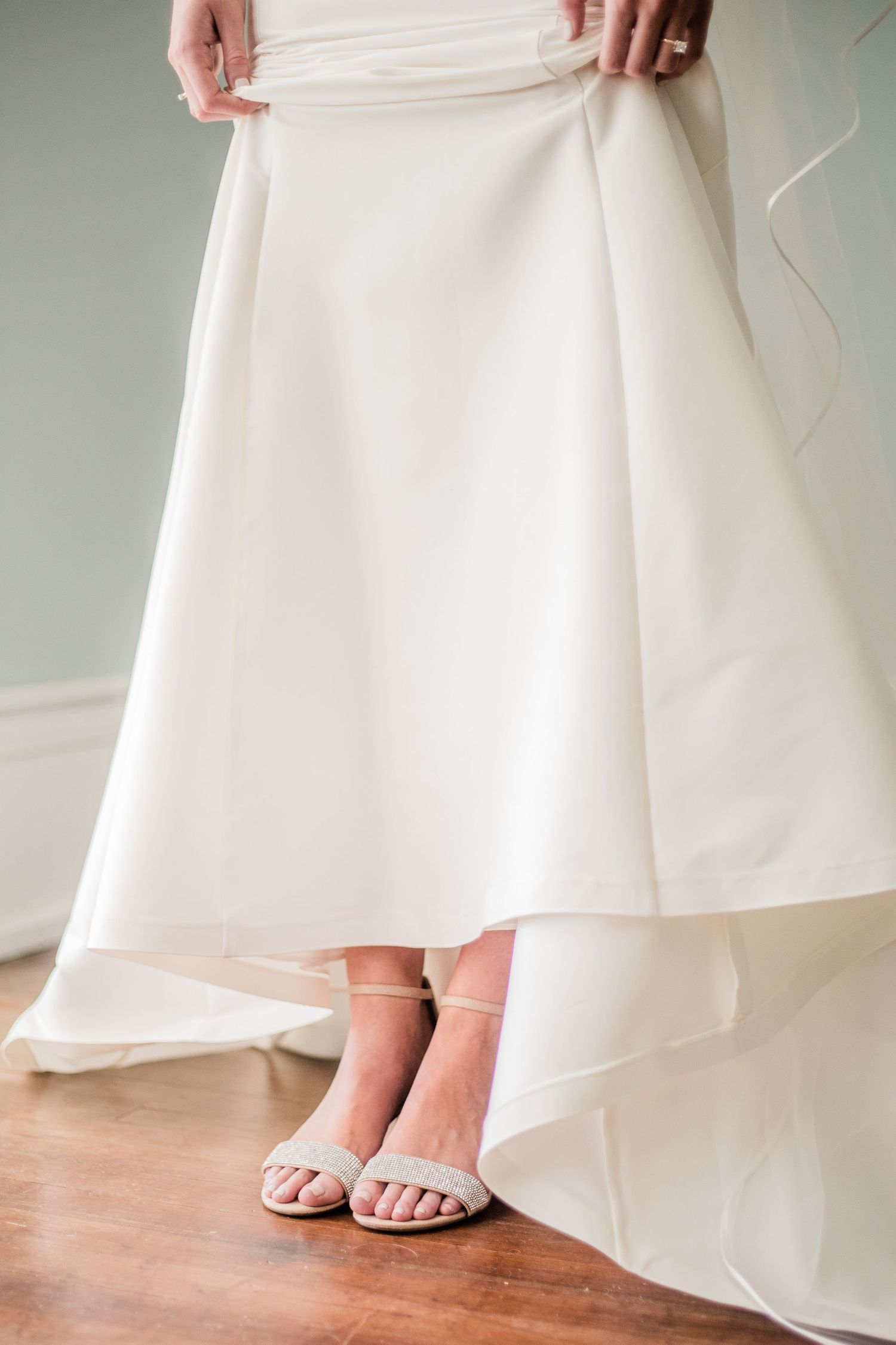 Close-up of white wedding dress hem and bridal shoes on hardwood floor.