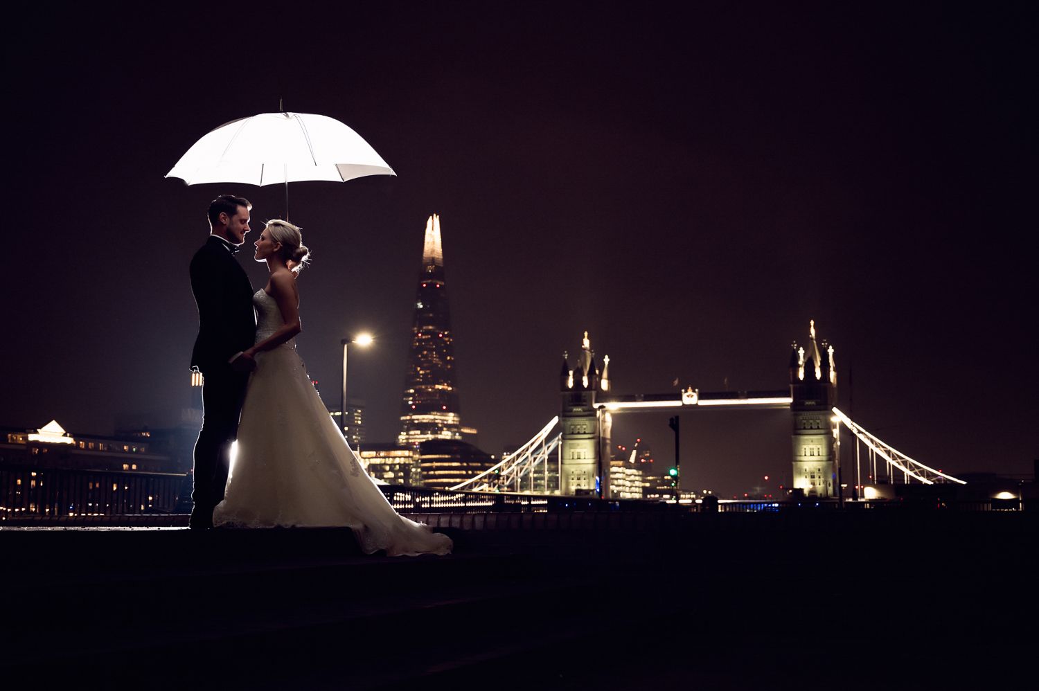 Silhouetted couple shares romantic moment under umbrella with London's Tower Bridge illuminated at night.