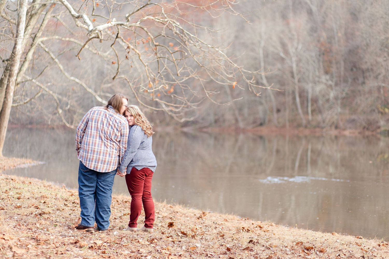 A couple walks along a lakeside path covered in fallen autumn leaves.