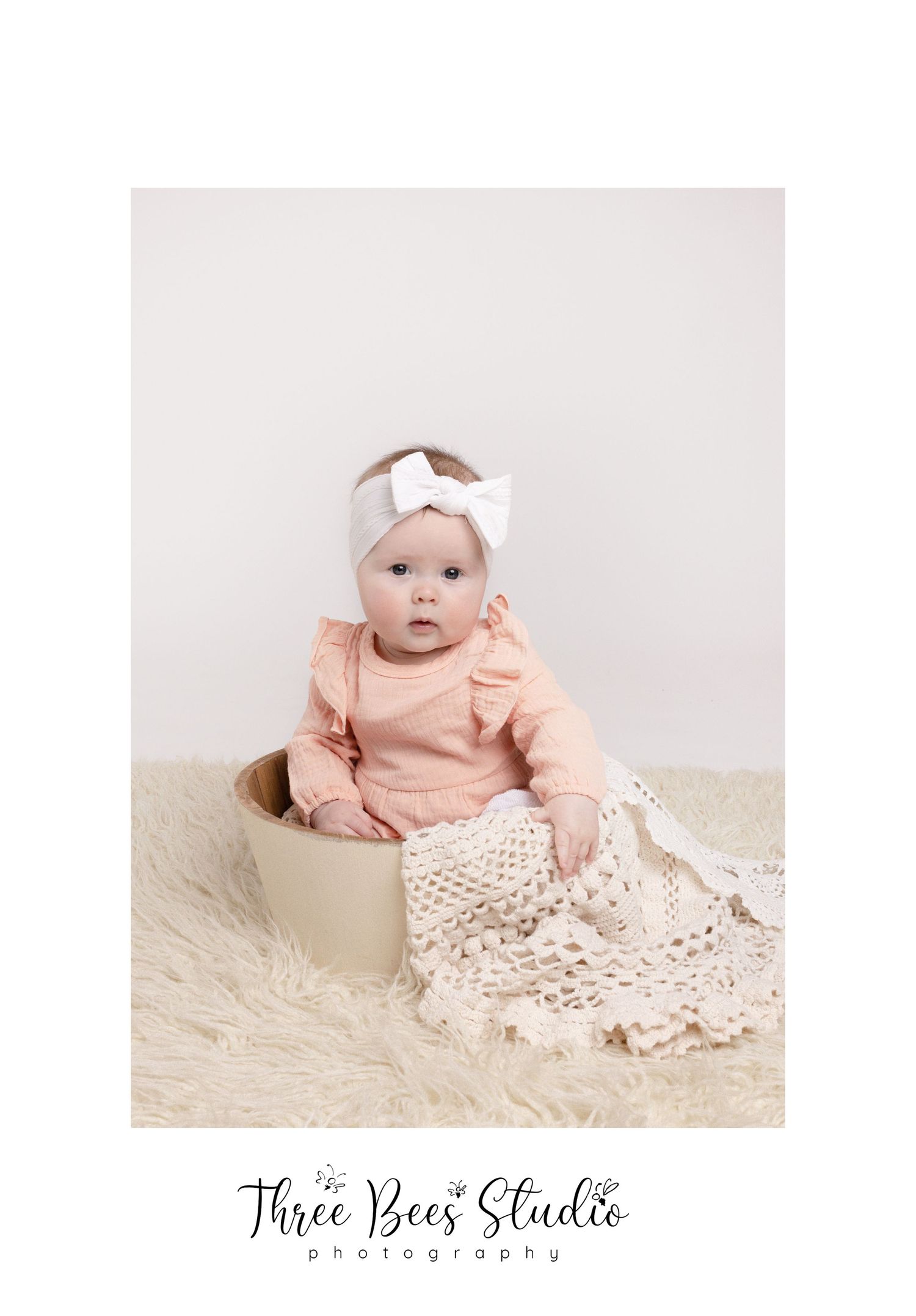Baby in peach outfit sitting in cream vessel with lace fabric during studio photo session.