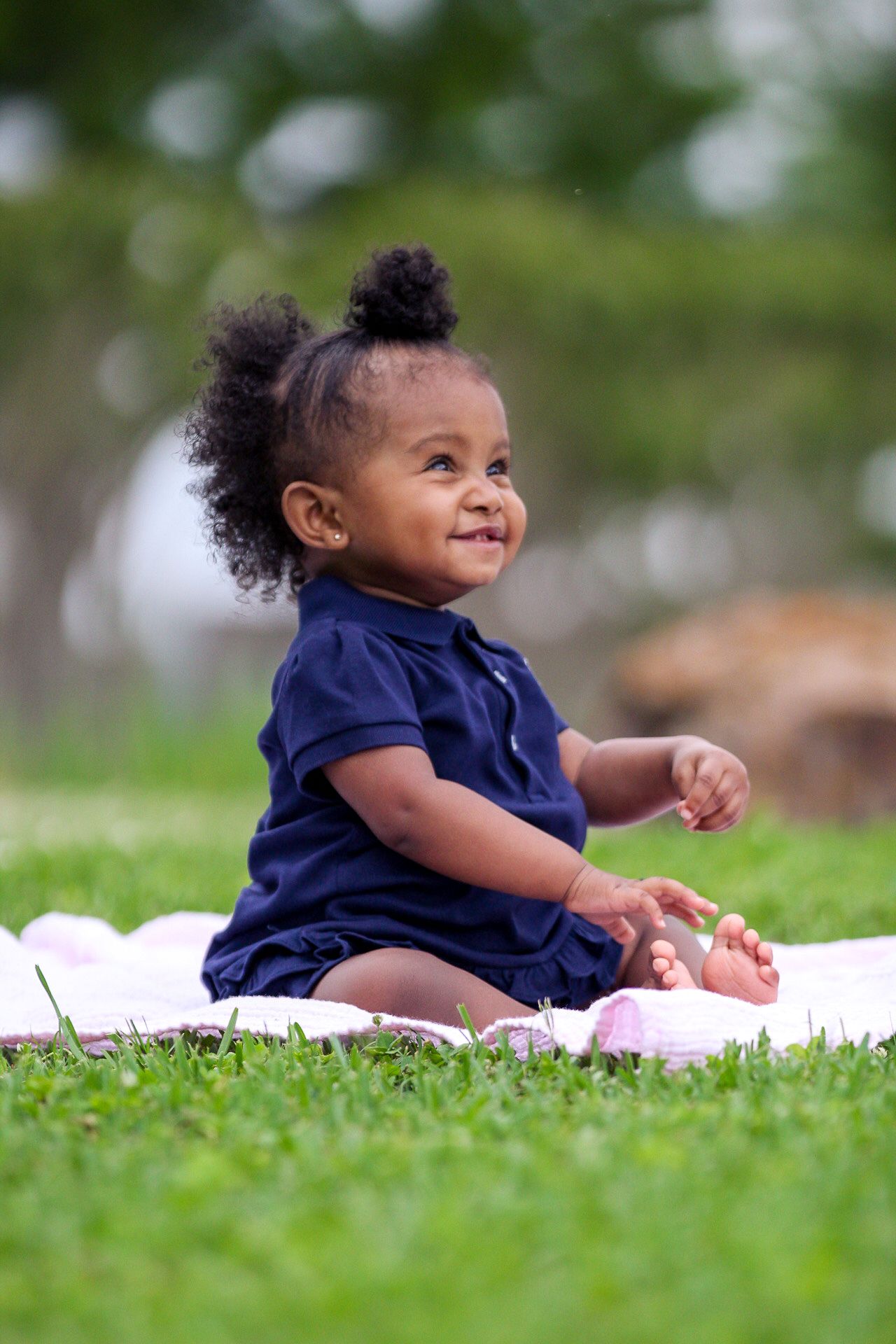 A baby in a navy blue shirt sits and smiles on a white blanket in the grass.