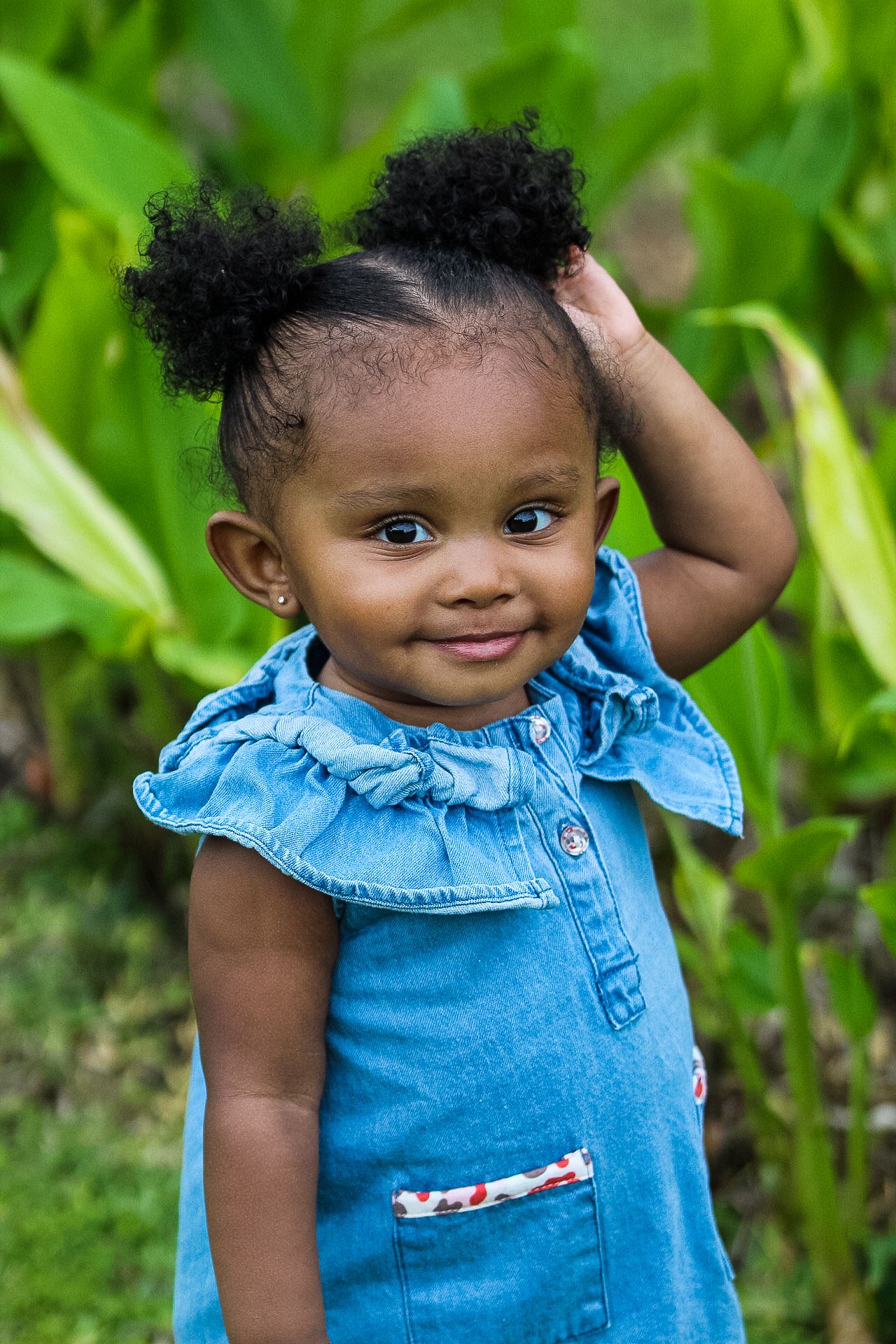 Toddler in denim dress touches hair while smiling in front of lush green foliage.