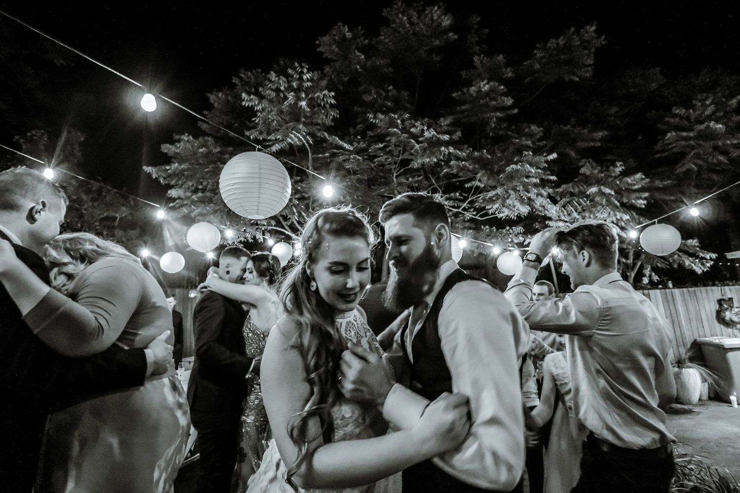 Wedding reception party outdoors at night with glowing paper lanterns and string lights creating a magical atmosphere.