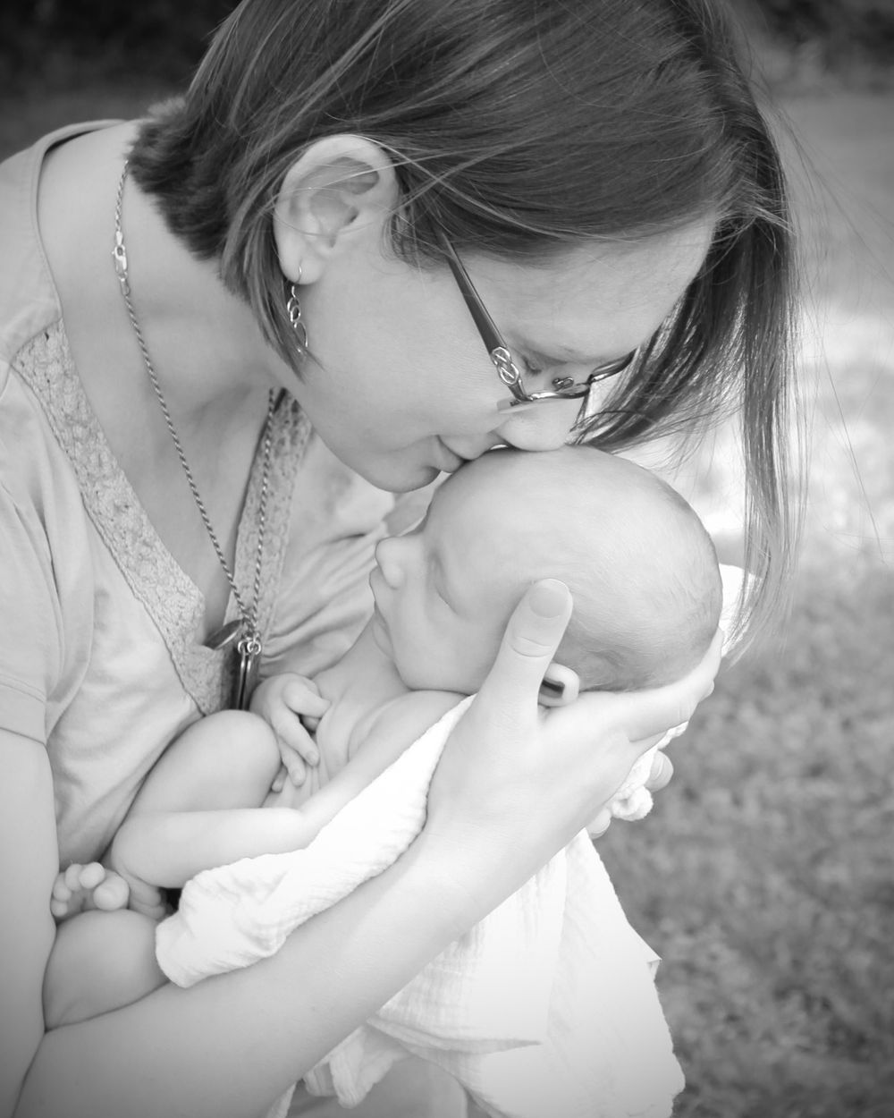 Black and white portrait of mother gently kissing newborn baby held in her arms outdoors.