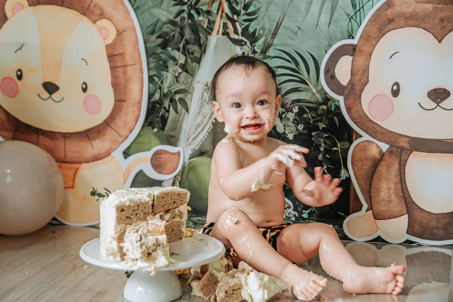 A jungle-themed cake smash photoshoot with safari animal decorations and balloons in front of a tropical backdrop.