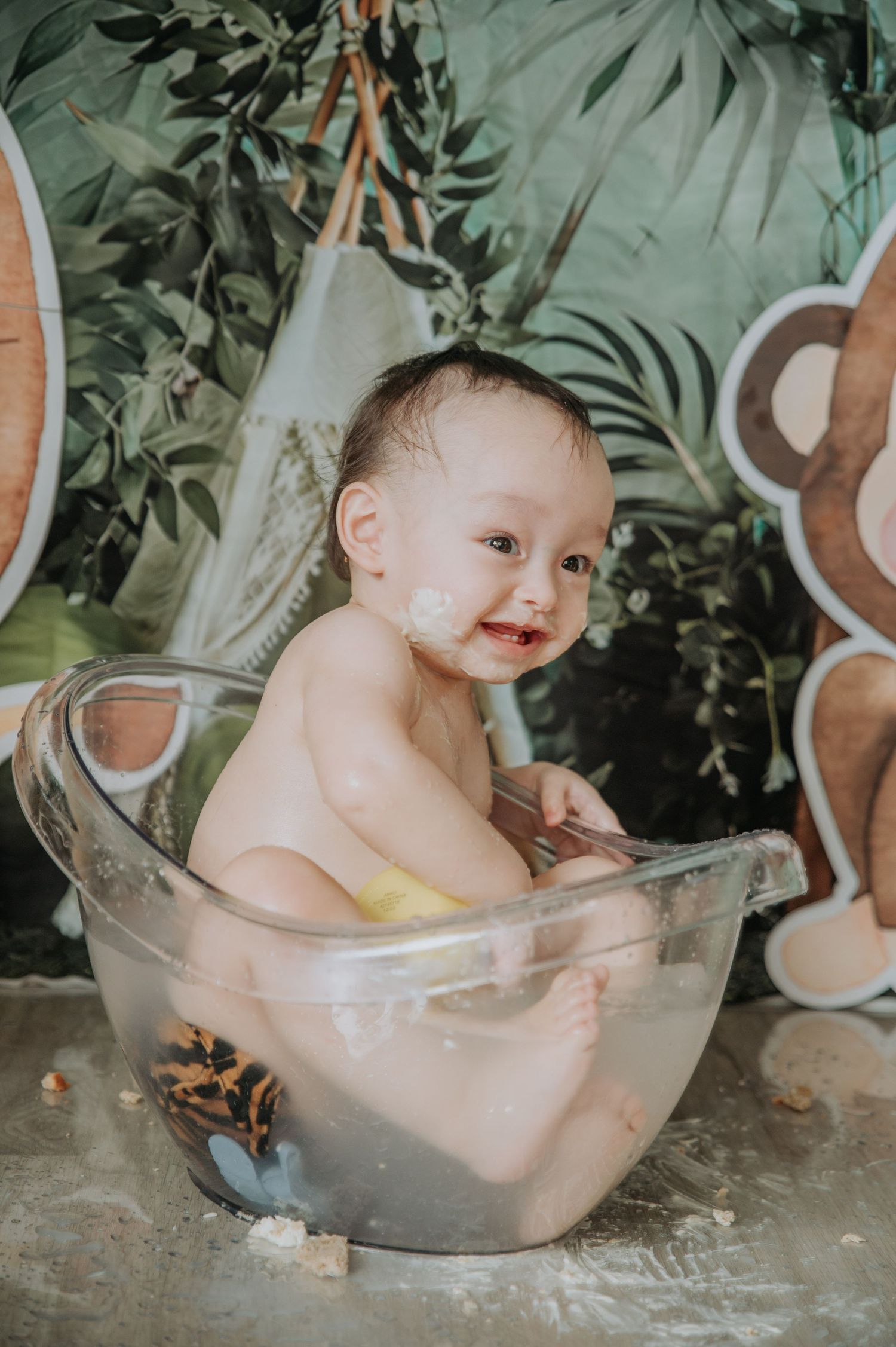 A festive cake smash photography setup with a clear bathtub prop against a tropical jungle-themed background.