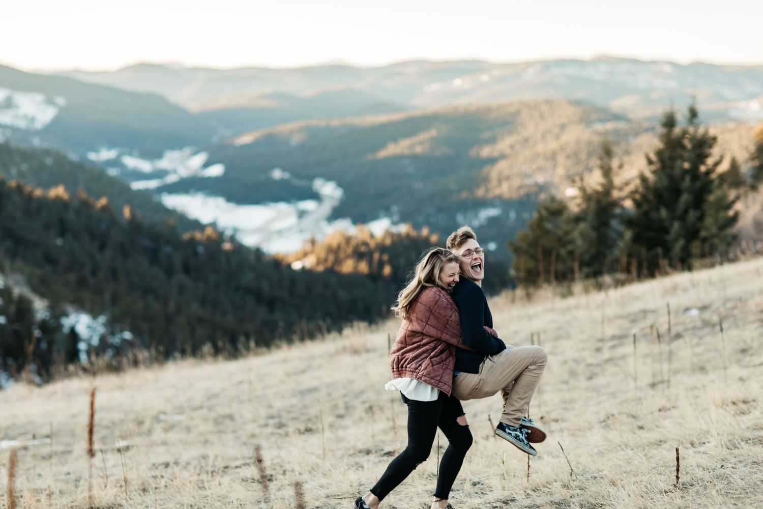 Girl in pink jacket picking boy in black sweater up while running in front of mountains with snow
