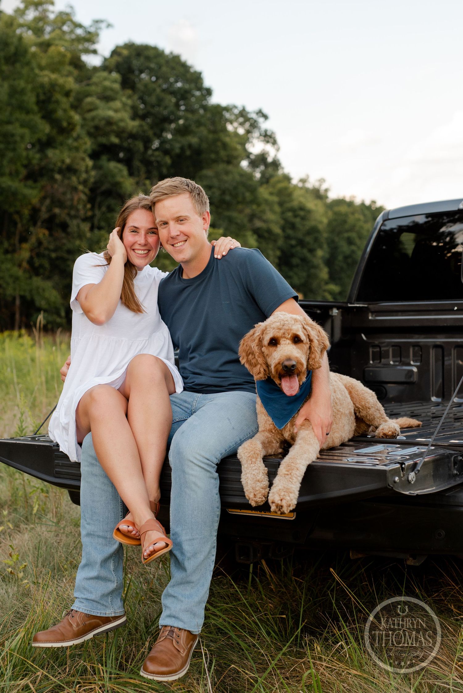 A couple and their goldendoodle relax together on the back of a black pickup truck in a summer field.