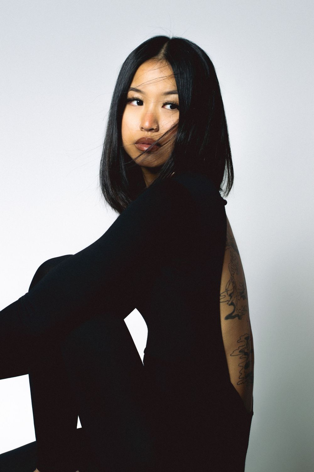 Artistic portrait in black attire with visible tattoo against white background.