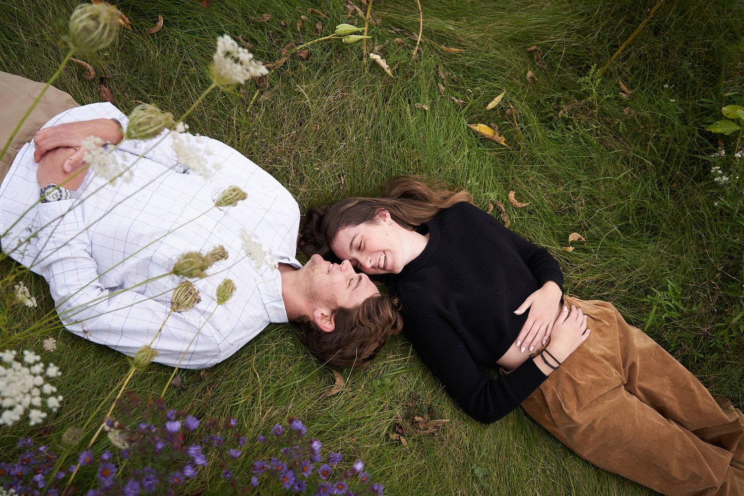 A couple lying on grass together surrounded by flowers and natural elements looking at each other and laughing.