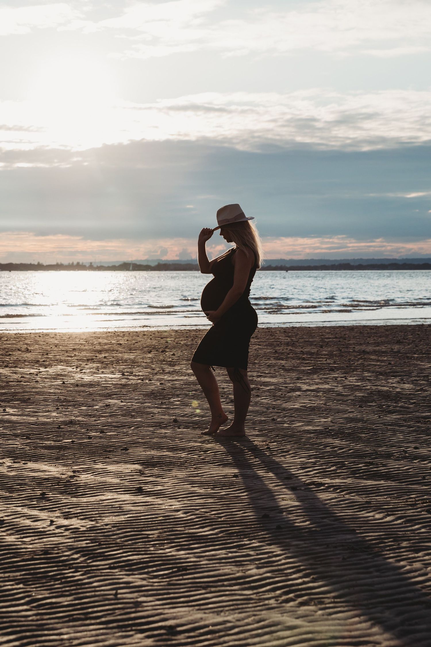 Silhouette of pregnant figure on beach at sunset casting long shadow on sand.
