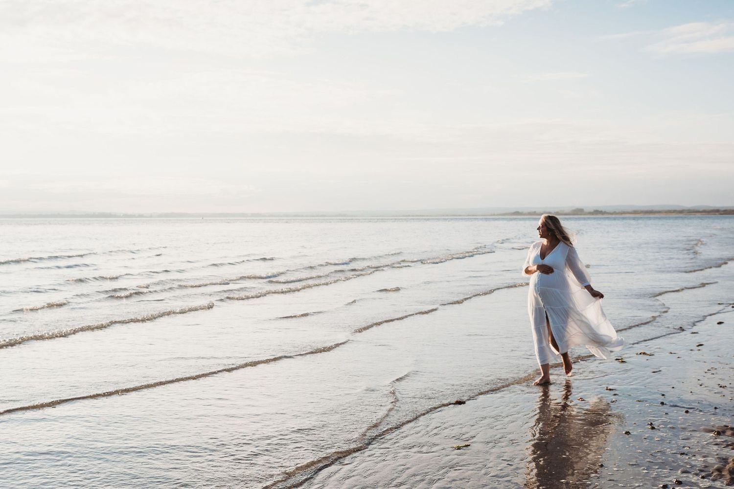 Figure in flowing white dress walking along beach shoreline at sunset.