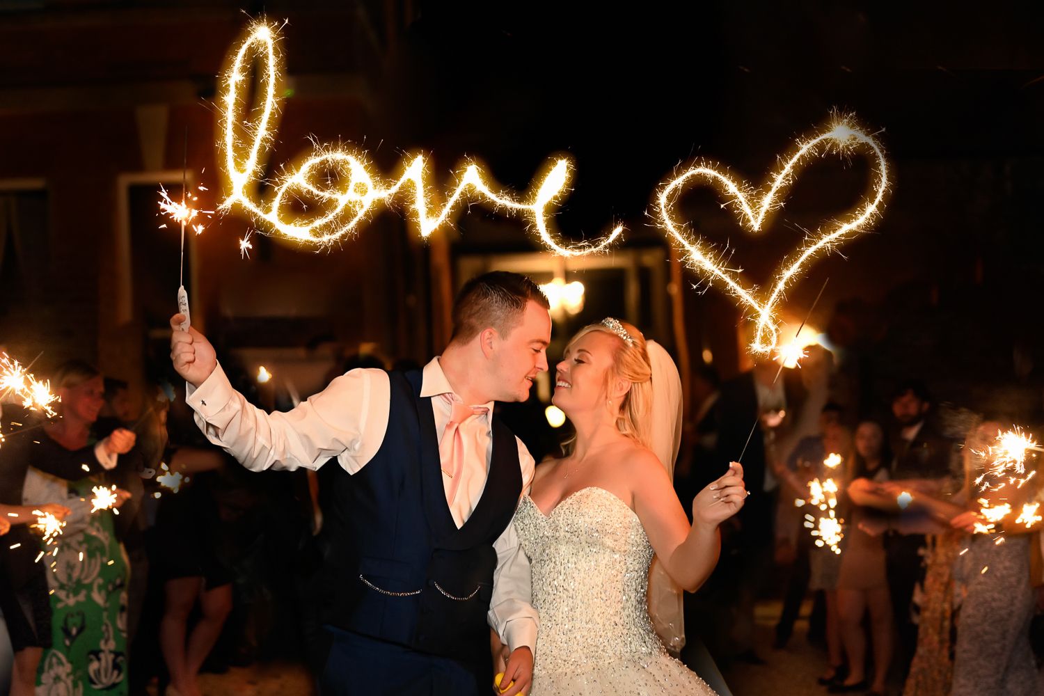 A sparkler creates a glowing love heart design behind a couple during their nighttime wedding celebration.