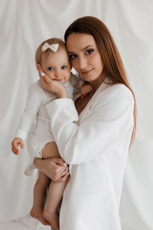 A heartwarming portrait of mother and baby in matching white outfits against a light background.