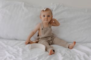 An infant in a beige knit romper sits on white bedding with a sweet expression.