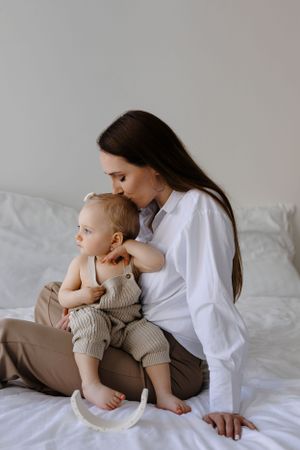 A tender moment between mother and small child sitting together on white bedding in casual attire.