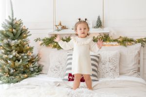 A toddler stands excitedly on a bed with Christmas decorations and tree in background.