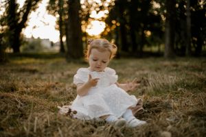 A toddler in a white lace dress sits thoughtfully on grass during golden hour.