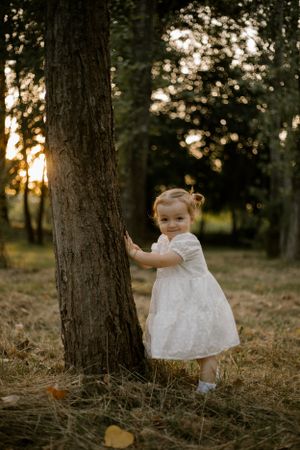 A small child in a white dress hugs a large tree trunk in a forest during sunset.