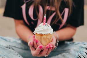 Hands holding a decorated cupcake with white frosting and sprinkles against denim.