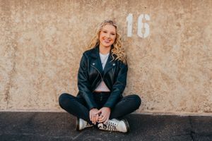 Teen in leather jacket sits cross-legged against concrete wall with number 16.
