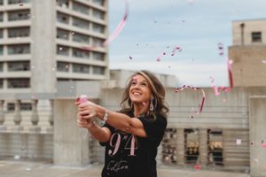 Person in black shirt celebrating with pink confetti against urban building backdrop.