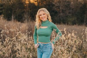 A green sweater and blue jeans create a casual autumn look against dried grasses and trees at golden hour.