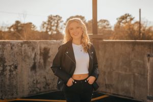 Teen in black leather jacket poses against industrial concrete wall at sunset.