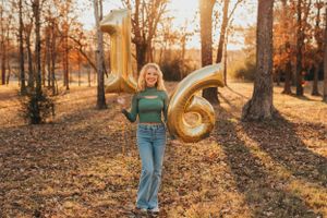 Birthday girl holds gold number balloons spelling 16 in autumn forest setting.